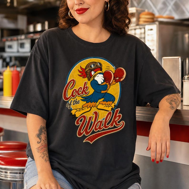 May include: A charcoal gray short-sleeve t-shirt featuring a graphic of a cartoon rooster wearing a brown aviator helmet and red boxing gloves. The design is set against a yellow circle and includes the text Cock of the Walk in red script.