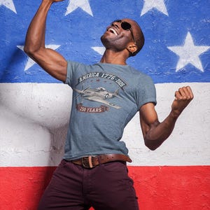 May include: A heather blue short-sleeve t-shirt featuring a graphic of a vintage fighter plane. The text reads AMERICA 1776-2026 and 250 YEARS. The design includes a small red star and a banner element, centered on the chest of the garment.