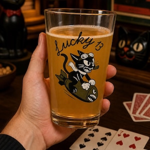 May include: A clear glass pint glass filled with a golden liquid, featuring a black graphic of a cartoon cat wearing goggles and a scarf while riding a green bomb. The text Lucky 13 is printed in black script above the cat illustration.