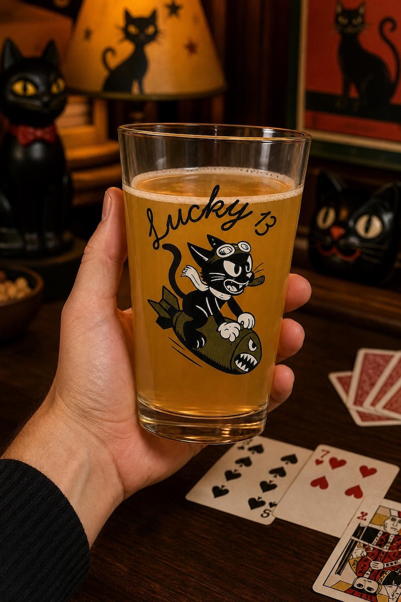 May include: A clear glass pint glass filled with a golden liquid, featuring a black graphic of a cartoon cat wearing goggles and a scarf while riding a green bomb. The text Lucky 13 is printed in black script above the cat illustration.