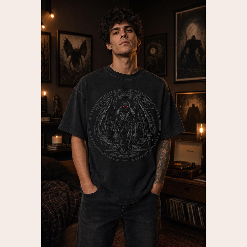 May include: A charcoal gray short-sleeve t-shirt featuring a circular graphic on the chest. The design depicts a winged creature with glowing red eyes, surrounded by the text Point Pleasant WV, 1966, and geographic coordinates in white.