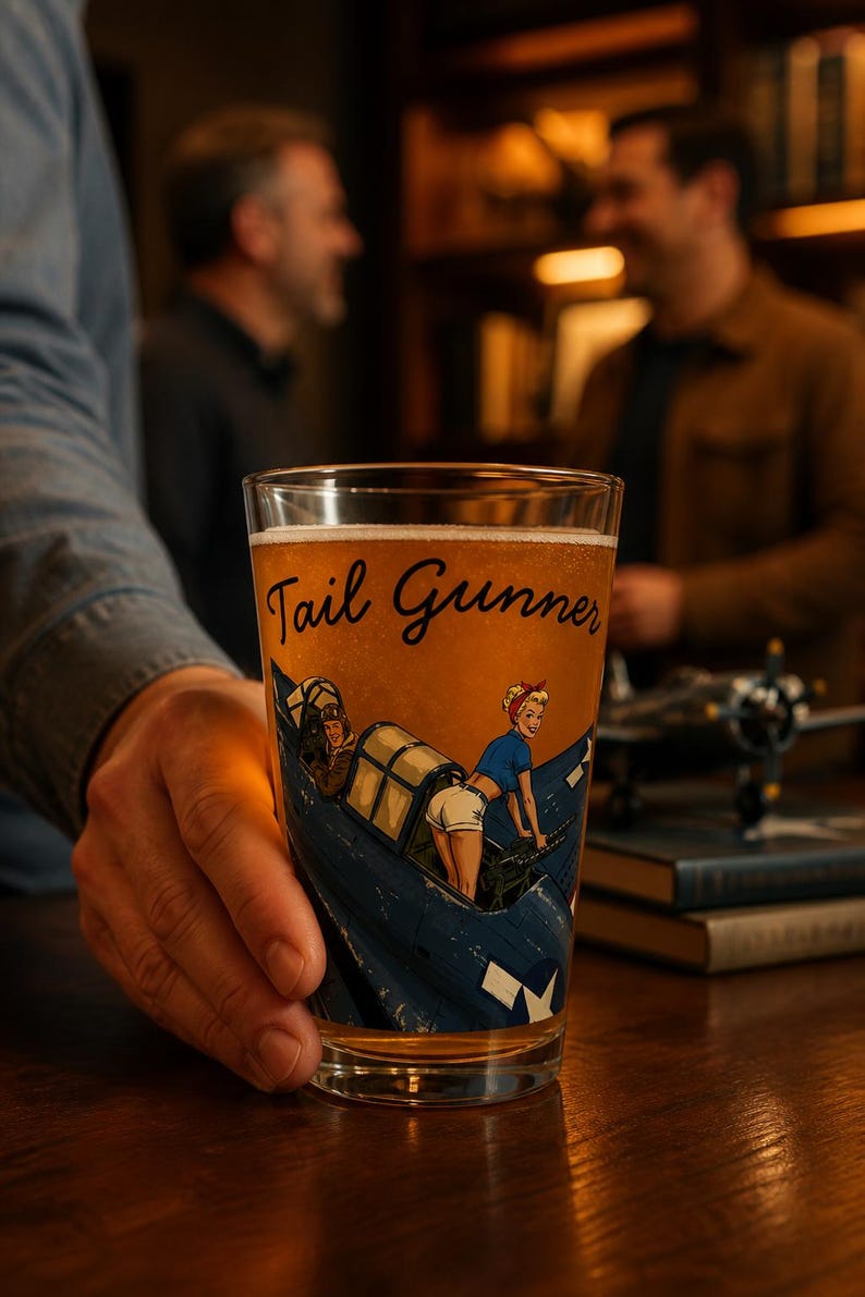 May include: A clear glass pint glass filled with amber beer, featuring a graphic illustration of a blue military aircraft and a person in a blue shirt and white shorts. The text Tail Gunner is printed in black script above the illustration.