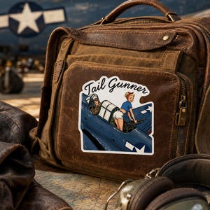 May include: A brown leather satchel featuring a sticker with the text Tail Gunner. The sticker depicts a blue military aircraft with a pilot and a person in a blue shirt and white shorts operating a machine gun. The bag has a top handle.