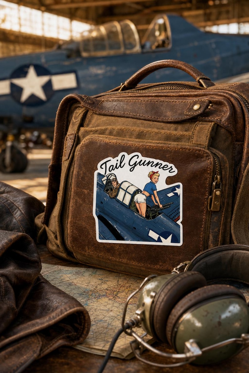 May include: A brown leather satchel featuring a sticker with the text Tail Gunner. The sticker depicts a blue military aircraft with a pilot and a person in a blue shirt and white shorts operating a machine gun. The bag has a top handle.