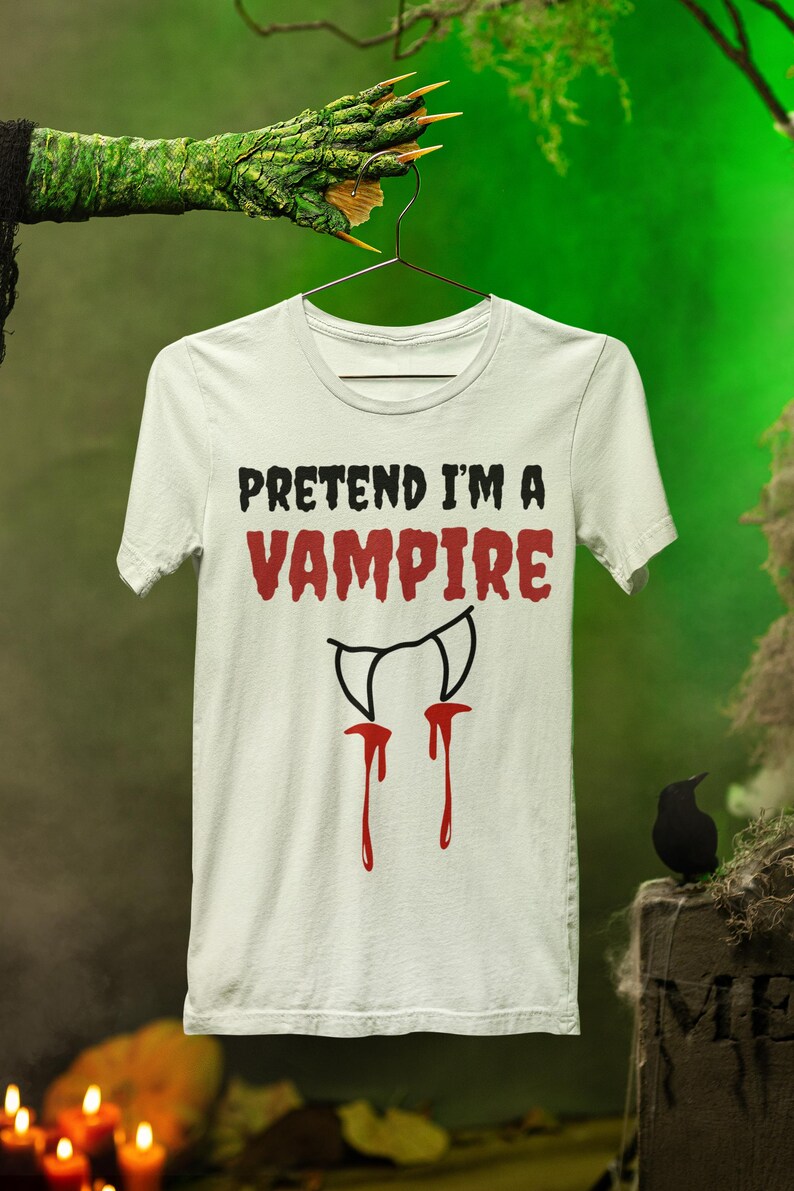 May include: White t-shirt with the text "PRETEND I'M A VAMPIRE" in black and red. Below the text is a graphic of vampire fangs with red blood dripping down. The shirt is hanging on a hanger against a green background.
