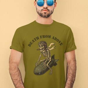 May include: Olive green t-shirt with the text "DEATH FROM ABOVE". The graphic shows a skeleton riding a bomb, wearing a pilot's helmet and scarf, and holding a cigar. The design is a vintage style.
