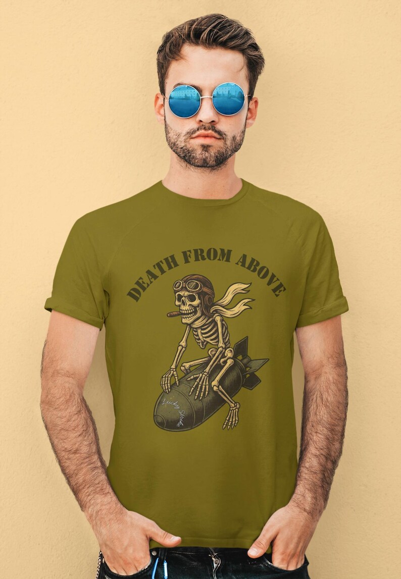 May include: Olive green t-shirt with the text "DEATH FROM ABOVE". The graphic shows a skeleton riding a bomb, wearing a pilot's helmet and scarf, and holding a cigar. The design is a vintage style.