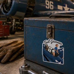May include: A blue metal toolbox featuring a sticker with the text Tail Gunner and an illustration of a person in a blue top and white shorts operating a machine gun in a cockpit. The box sits near brown leather gloves and a metal ratchet tool.
