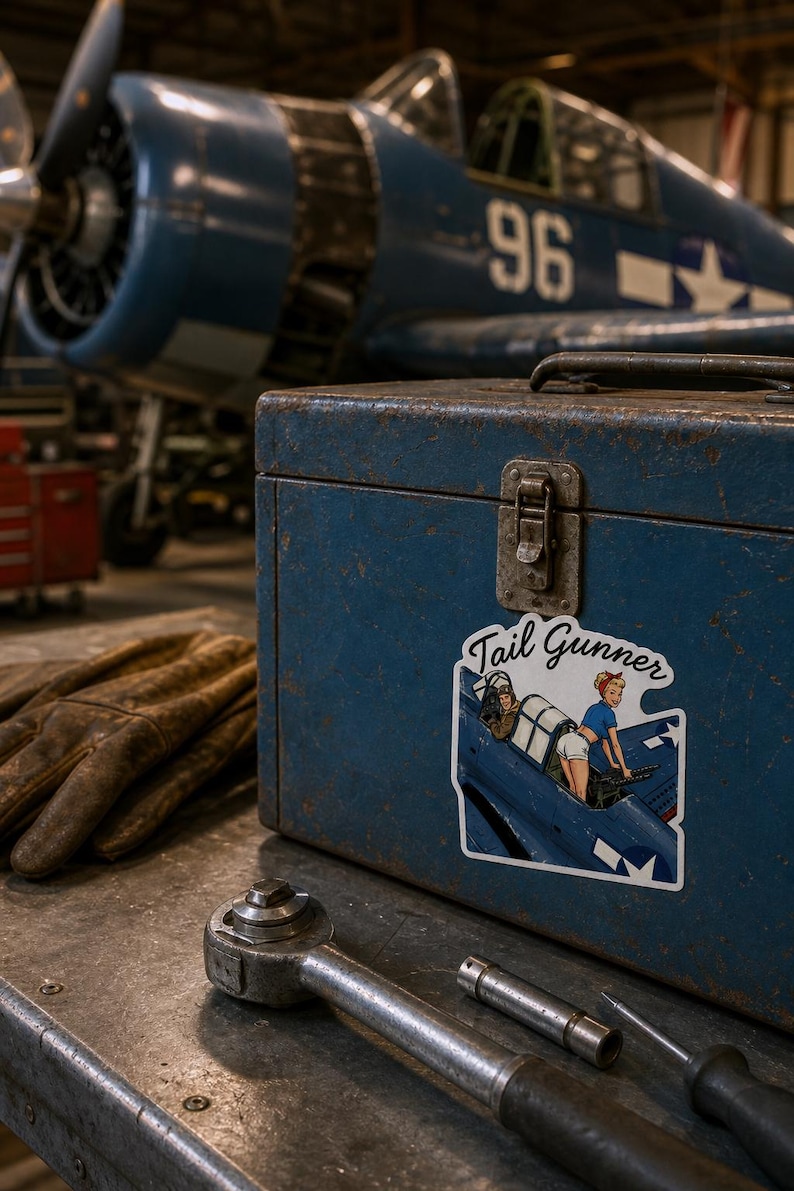 May include: A blue metal toolbox featuring a sticker with the text Tail Gunner and an illustration of a person in a blue top and white shorts operating a machine gun in a cockpit. The box sits near brown leather gloves and a metal ratchet tool.