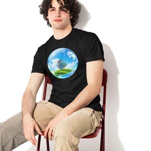 May include: A black short-sleeve t-shirt featuring a circular graphic print of a blue discus fish swimming inside a transparent glass bubble that contains a small green grassy island under a bright blue sky with white clouds.