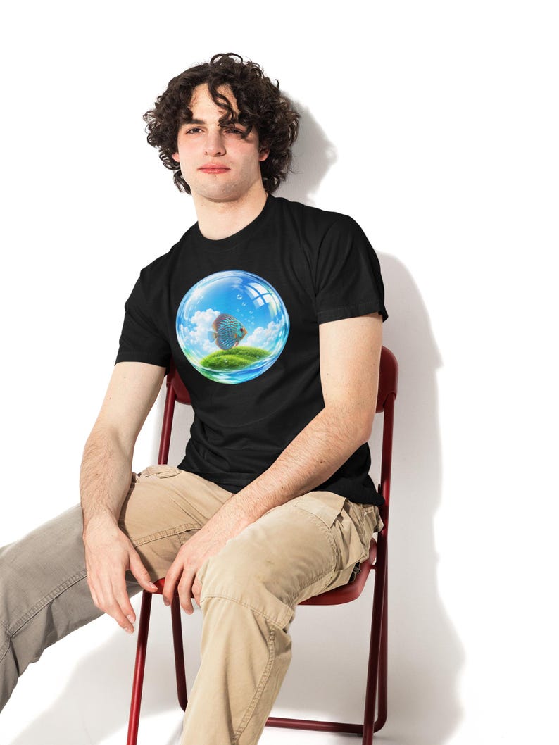 May include: A black short-sleeve t-shirt featuring a circular graphic print of a blue discus fish swimming inside a transparent glass bubble that contains a small green grassy island under a bright blue sky with white clouds.