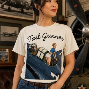 May include: An off-white short-sleeve t-shirt featuring a graphic print of a blue military aircraft with a pilot and a pin-up style figure operating a machine gun. The text "Tail Gunner" is printed in black script above the illustration.