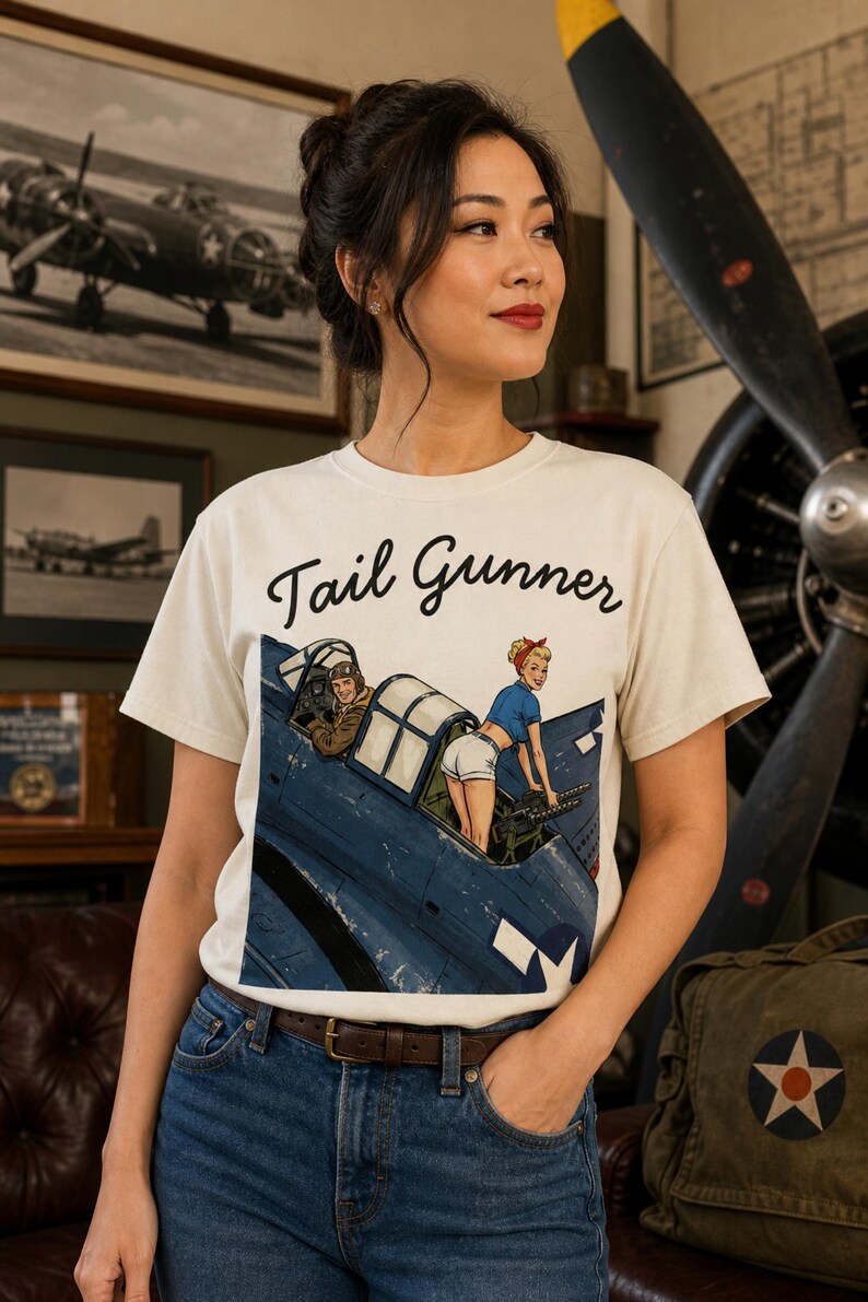 May include: An off-white short-sleeve t-shirt featuring a graphic print of a blue military aircraft with a pilot and a pin-up style figure operating a machine gun. The text "Tail Gunner" is printed in black script above the illustration.