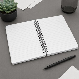 May include: An open spiral-bound notebook with white lined pages and a black metal coil spine. The notebook is positioned on a flat surface alongside a sleek black ballpoint pen, a small potted succulent plant, and a dark glass container.