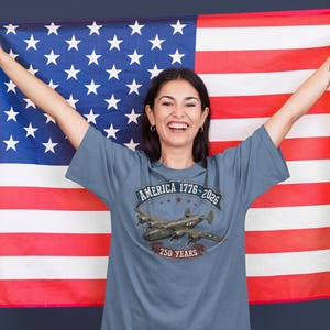 May include: A slate blue t-shirt featuring a graphic print of a vintage military aircraft. The text reads AMERICA 1776-2026 and 250 YEARS. The shirt is displayed against a large American flag with red, white, and blue stripes and white stars.