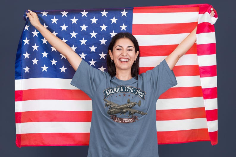 May include: A slate blue t-shirt featuring a graphic print of a vintage military aircraft. The text reads AMERICA 1776-2026 and 250 YEARS. The shirt is displayed against a large American flag with red, white, and blue stripes and white stars.