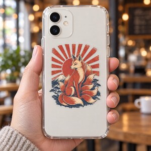 May include: A clear smartphone case featuring a graphic illustration of a multi-tailed fox sitting in front of a red sunburst, a mountain peak, and stylized trees. The design is centered on the back of the transparent protective cover.
