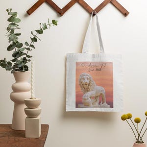 May include: A white tote bag featuring a lion statue image with the text "St. Augustine FL Est. 1565". The bag hangs from a wooden wall rack. A pink vase and a candle holder are on a wooden table. The image has a sunset background.