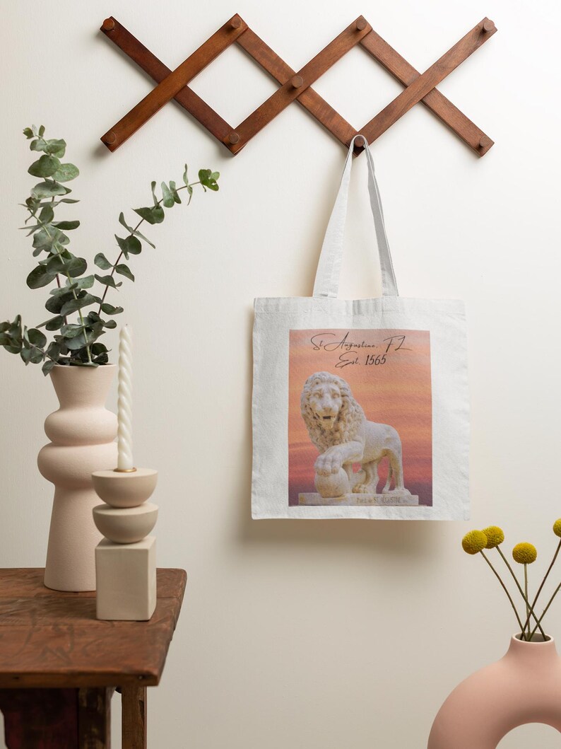 May include: A white tote bag featuring a lion statue image with the text "St. Augustine FL Est. 1565". The bag hangs from a wooden wall rack. A pink vase and a candle holder are on a wooden table. The image has a sunset background.