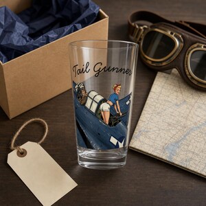May include: A clear glass tumbler featuring a printed graphic of a blue military aircraft with a person in a blue shirt and white shorts positioned in the cockpit. The text Tail Gunner is printed in black script above the aircraft illustration.