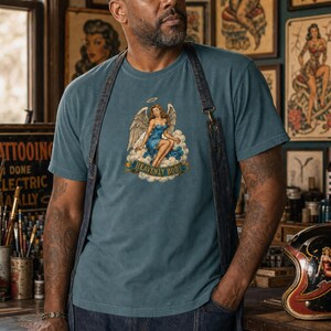 May include: A blue t-shirt featuring a graphic print of an angel with wings sitting on a cloud, wearing a blue dress, with a halo above her head and a banner below the cloud that reads "HEAVENLY BODY" in gold lettering on a blue background.