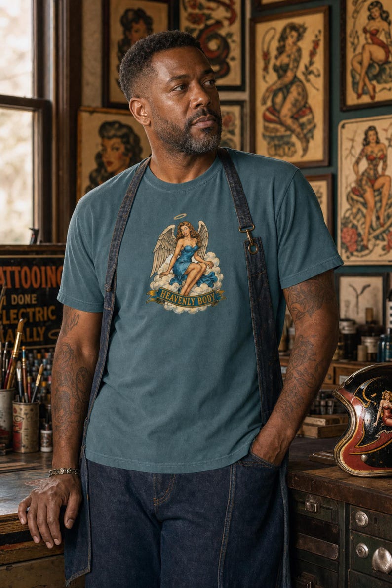 May include: A blue t-shirt featuring a graphic print of an angel with wings sitting on a cloud, wearing a blue dress, with a halo above her head and a banner below the cloud that reads "HEAVENLY BODY" in gold lettering on a blue background.