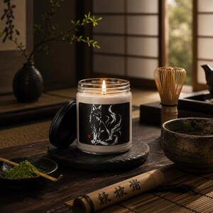 May include: A clear glass jar candle with a lit wick and a black label featuring a white illustration of a fox with a flowing tail. The candle has a black lid resting beside it on a dark stone coaster, set among various tea-related items.