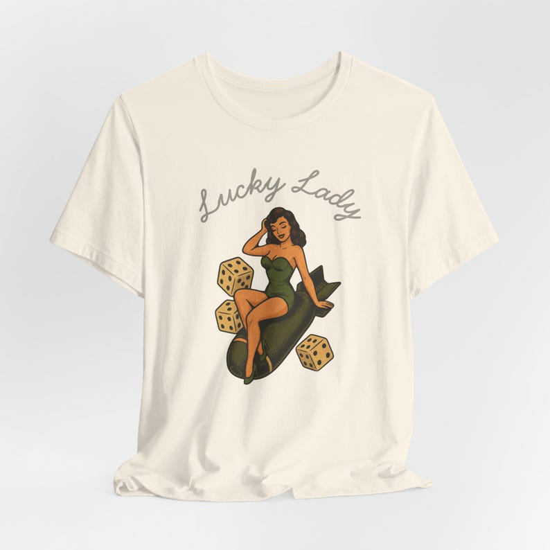 May include: A cream-colored short-sleeve t-shirt featuring a graphic illustration of a person in a green swimsuit sitting on a dark bomb, surrounded by three dice. The text Lucky Lady is printed in a cursive font above the central illustration.