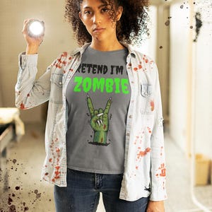May include: A gray t-shirt with the text "PRETEND I'M ZOMBIE" in black and green, featuring a green zombie hand making a rock and roll gesture. The person is wearing a light blue denim shirt and blue jeans.