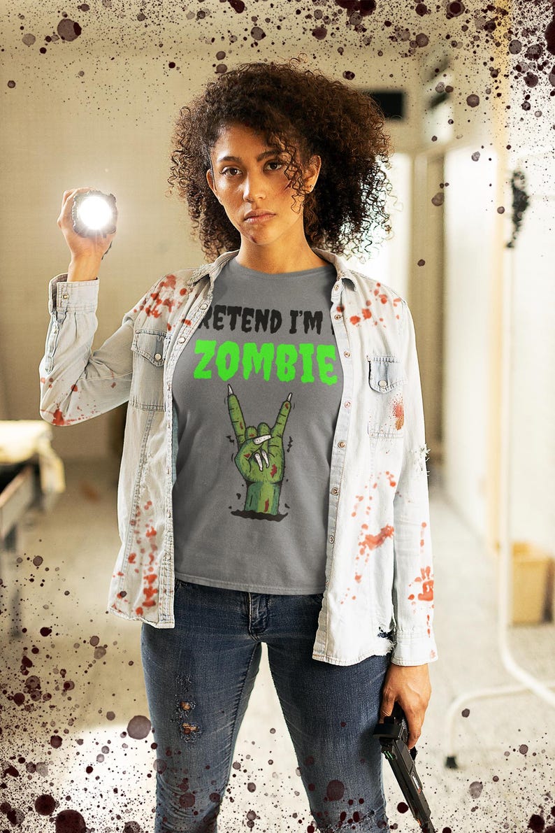 May include: A gray t-shirt with the text "PRETEND I'M ZOMBIE" in black and green, featuring a green zombie hand making a rock and roll gesture. The person is wearing a light blue denim shirt and blue jeans.