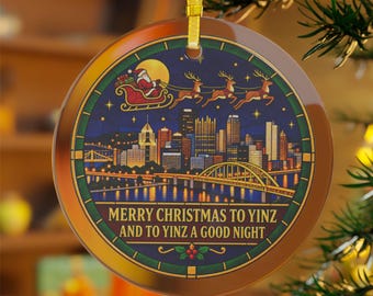 May include: A round ceramic Christmas ornament featuring a stained-glass style illustration of the Pittsburgh skyline at night with Santa in a sleigh above. The text reads MERRY CHRISTMAS TO YINZ AND TO YINZ A GOOD NIGHT in gold lettering.