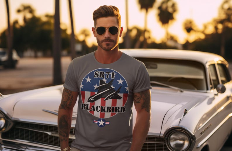 May include: Gray t-shirt featuring a graphic of the SR-71 Blackbird aircraft within a patriotic design of stars and stripes. The text "SR71 BLACKBIRD" is displayed on the shirt. The person is wearing sunglasses and standing in front of a classic white car.