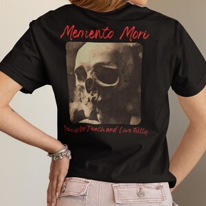 May include: Black t-shirt with a skull graphic and the words "Memento Mori" in red script. The phrase "Remember Death and Live Fully" is below the skull image. The shirt is a classic crew neck.