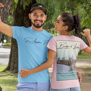 Peut inclure: Deux personnes portant des t-shirts. L'un est bleu clair avec "St. Augustine, FL" en &eacute;criture cursive. L'autre est rose avec un graphique du pont des Lions et le texte "Where Lions Guard the Saint Augustine, FL".