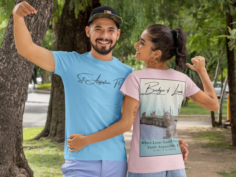 Peut inclure: Deux personnes portant des t-shirts. L'un est bleu clair avec "St. Augustine, FL" en &eacute;criture cursive. L'autre est rose avec un graphique du pont des Lions et le texte "Where Lions Guard the Saint Augustine, FL".