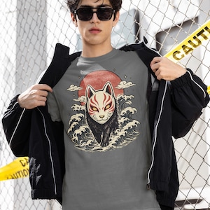 May include: A grey t-shirt featuring a graphic illustration of a black fox wearing a white and red mask, positioned in front of a large red sun and stylised ocean waves. The design includes small star accents and clouds surrounding the fox figure.