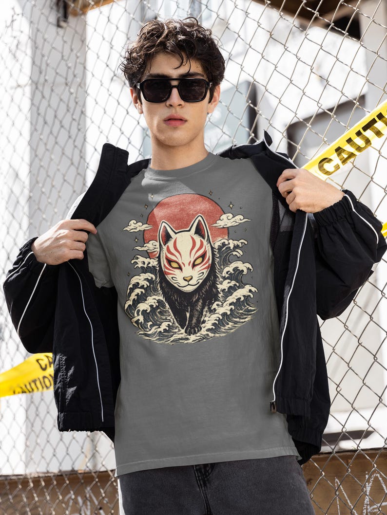 May include: A grey t-shirt featuring a graphic illustration of a black fox wearing a white and red mask, positioned in front of a large red sun and stylised ocean waves. The design includes small star accents and clouds surrounding the fox figure.