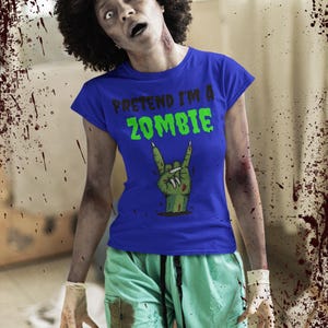 May include: A person in a blue t-shirt with the text "PRETEND I'M A ZOMBIE" in green. The shirt features a zombie hand graphic. The person is wearing green scrubs and has zombie makeup.