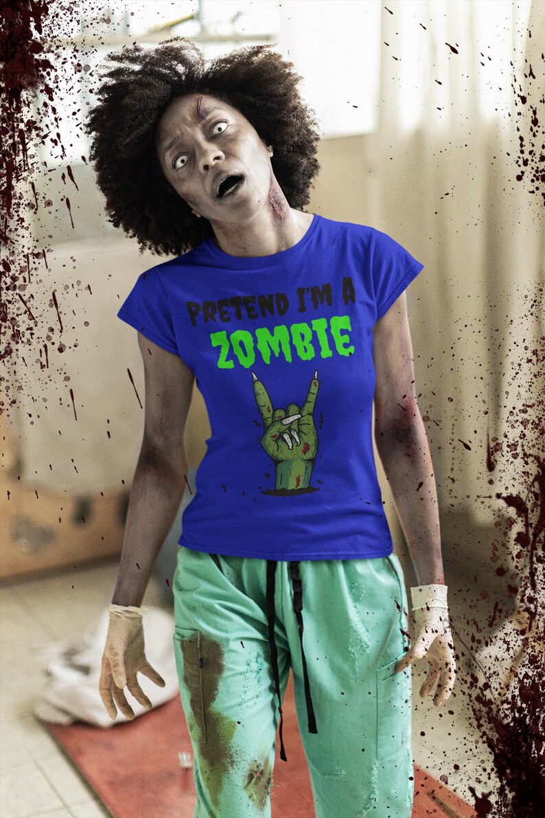 May include: A person in a blue t-shirt with the text "PRETEND I'M A ZOMBIE" in green. The shirt features a zombie hand graphic. The person is wearing green scrubs and has zombie makeup.
