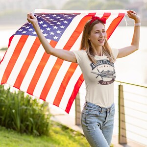 May include: A cream-colored short-sleeve t-shirt featuring a graphic of a vintage propeller airplane, the text AMERICA 1776-2026, and the words 250 YEARS. The shirt is paired with blue denim jeans and a red hair accessory, held with a large American flag.