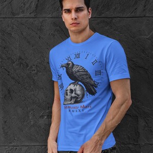 May include: A light blue t-shirt featuring a graphic of a raven perched on a skull, set against a clock face. The text "Memento Mori" is below the skull. The shirt is worn by a person.