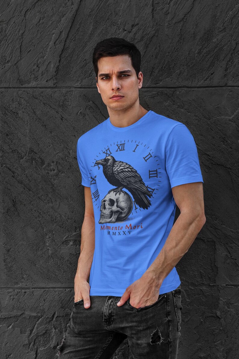 May include: A light blue t-shirt featuring a graphic of a raven perched on a skull, set against a clock face. The text "Memento Mori" is below the skull. The shirt is worn by a person.