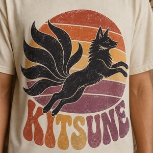 May include: A cream-colored short-sleeve t-shirt featuring a graphic of a black nine-tailed fox leaping in front of a distressed sunset circle. The word KITSUNE is printed in large, wavy, multi-colored retro-style lettering below the fox.