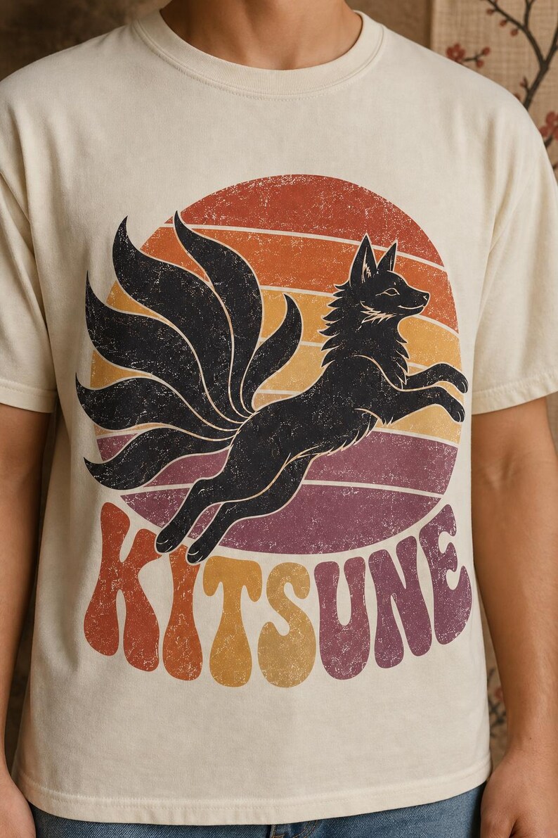 May include: A cream-colored short-sleeve t-shirt featuring a graphic of a black nine-tailed fox leaping in front of a distressed sunset circle. The word KITSUNE is printed in large, wavy, multi-colored retro-style lettering below the fox.