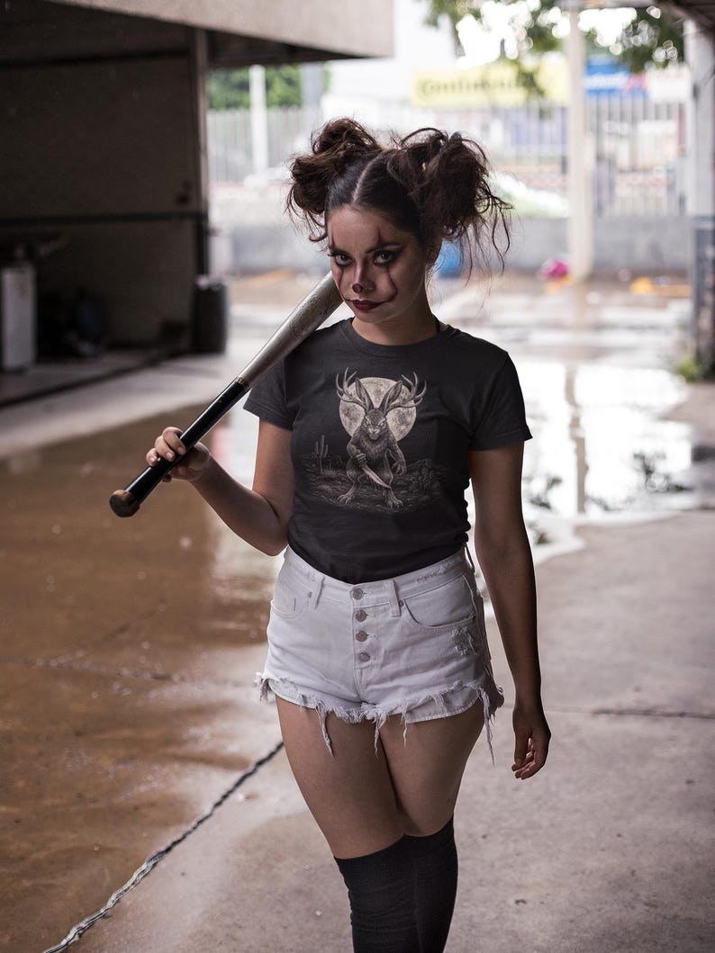 K&ouml;nnte beinhalten: Ein dunkelgraues Kurzarm-T-Shirt mit einem Grafikdruck eines Jackalopen, der ein Messer in einer W&uuml;stenlandschaft vor einem Vollmond h&auml;lt. Dazu werden wei&szlig;e Jeansshorts mit ausgefranstem Saum und schwarze Overknee-Socken getragen.
