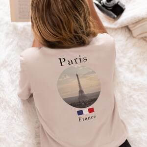 May include: A light pink t-shirt featuring the Eiffel Tower in a circular design, with the word "Paris" above and "France" below. The French flag is also displayed. The shirt is worn by a person.