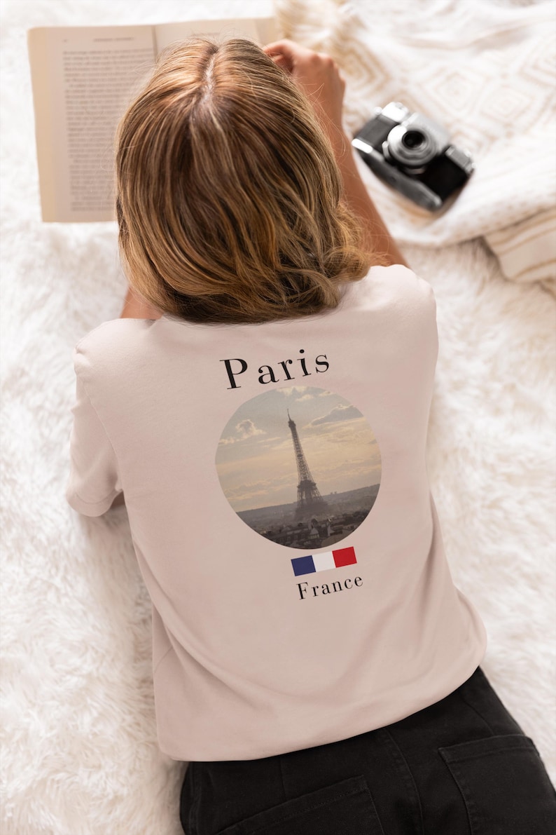 May include: A light pink t-shirt featuring the Eiffel Tower in a circular design, with the word "Paris" above and "France" below. The French flag is also displayed. The shirt is worn by a person.