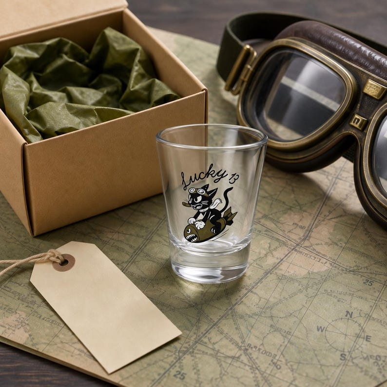 May include: A clear glass shot glass featuring a black graphic of a cartoon cat wearing goggles and riding an olive green bomb, with the text Lucky 13 printed above the cat. The glass is positioned near a brown cardboard box and aviator goggles.