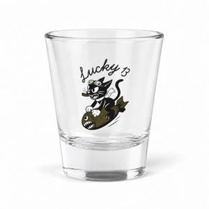 May include: A clear glass shot glass featuring a black and white cartoon illustration of a cat wearing goggles and riding a bomb. Above the cat, the text Lucky 13 is printed in a black script font. The bomb has a shark-like mouth design.