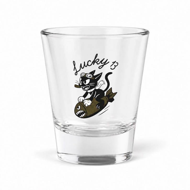 May include: A clear glass shot glass featuring a black and white cartoon illustration of a cat wearing goggles and riding a bomb. Above the cat, the text Lucky 13 is printed in a black script font. The bomb has a shark-like mouth design.